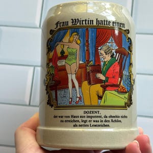 May include: A beige ceramic beer stein featuring a colorful illustration of a woman in a green corset and a man in a red jacket reading a book titled Achilos. The text reads Frau Wirtin hatte einen DOZENT, der war von Haus aus impotent, da ohnehin nichts zu erreichen, legt er was in den Ächilos, als nettes Lesezeichen.
