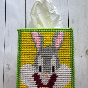 May include: A handmade tissue box cover featuring a cartoon rabbit design. The cover is made of yarn in yellow, gray, pink, white, and green. The rabbit's face is in the center, with a tissue emerging from the top.