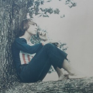 May include: A person with long, brown hair sits on a tree branch, wearing a blue shirt with red and white stripes and blue jeans. The person is looking off to the side, with a thoughtful expression on their face.
