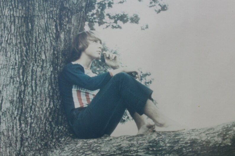 May include: A person with long, brown hair sits on a tree branch, wearing a blue shirt with red and white stripes and blue jeans. The person is looking off to the side, with a thoughtful expression on their face.