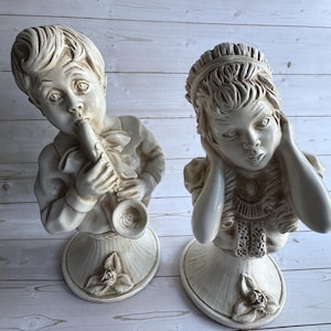 Puede incluir: Dos bustos de cerámica blanquecina. Uno representa a un niño tocando un instrumento de viento, el otro a una niña tapándose los oídos. Ambos están sobre bases ovaladas con detalles florales, sugiriendo un estilo vintage o antiguo.