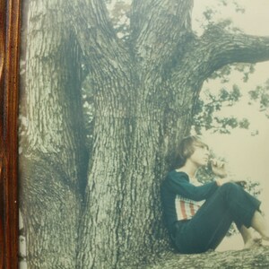 May include: A vintage photograph of a woman sitting on a tree branch. The woman is wearing a dark blue dress and has her hair pulled back. The tree is large and has a thick trunk. The photo is in a wooden frame.