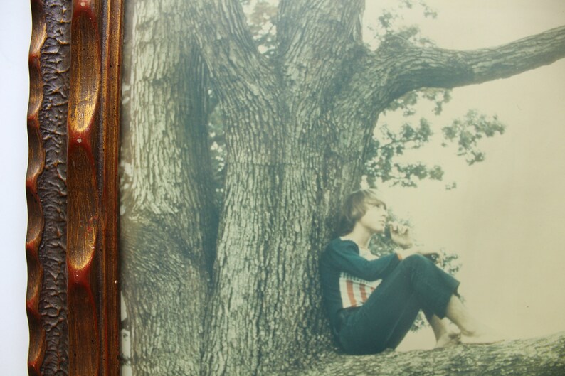 May include: A vintage photograph of a woman sitting on a tree branch. The woman is wearing a dark blue dress and has her hair pulled back. The tree is large and has a thick trunk. The photo is in a wooden frame.