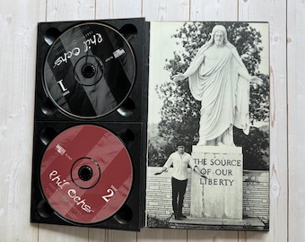 Puede incluir: Una caja de CD negra abierta que muestra dos discos y una imagen impresa. Los discos están marcados con "1" y "2" con el nombre del artista. La imagen muestra una estatua y el texto "THE SOURCE OF OUR LIBERTY."