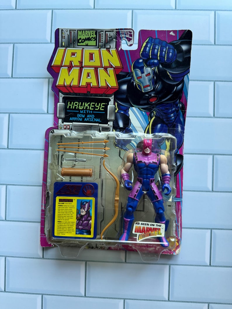 May include: A Hawkeye action figure from Marvel Comics, packaged on a colorful card. The figure is in a dynamic pose, with a bow and arrow. The packaging features the Iron Man logo and the text "Hawkeye with Bow and Arrow Arsenal."