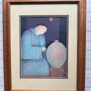 May include: Framed art print featuring a figure in a blue patterned robe, with a large, light-colored vase. A red circle is in the upper portion of the image. The artwork is framed in a brown wooden frame with a cream-colored mat.