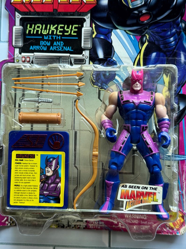 May include: Hawkeye action figure in original packaging. The figure is in a blue and purple costume, with a bow and arrow arsenal. The packaging features the text "Hawkeye with Bow and Arrow Arsenal" and "As Seen on the Marvel Action Hour".