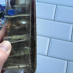 May include: A clear plastic package, possibly for a toy or collectible, held by a hand. The package has multiple compartments and a partially visible product inside. The background is a light blue tiled wall.