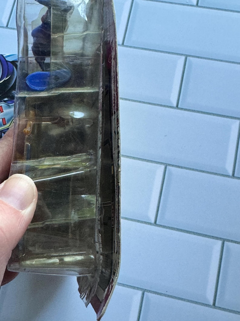 May include: A clear plastic package, possibly for a toy or collectible, held by a hand. The package has multiple compartments and a partially visible product inside. The background is a light blue tiled wall.