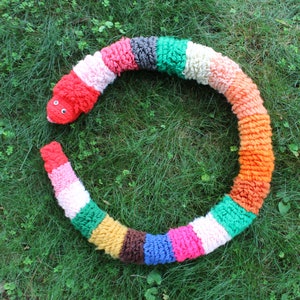 May include: A colorful crocheted snake toy with a red head and a friendly face. The snake is made of different colored yarn and is curled into a circle.
