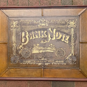Op de afbeelding: Een ingelijst, rechthoekig kunstwerk met een bruin, leerachtig oppervlak. Het kunstwerk toont de woorden "BANK NOTE" in grote, sierlijke letters. Het ontwerp bevat goudkleurige accenten en ingewikkelde details, die lijken op een antieke bankbiljet.