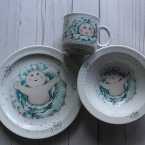 Puede incluir: Un juego de vajilla infantil de cerámica blanca con un diseño de Cabbage Patch Kid. El juego incluye un tazón, un plato y una taza. Cada pieza presenta una imagen de dibujos animados de un bebé en una col.