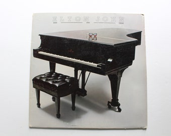 May include: A black grand piano with a black piano bench. The piano has a white keyboard and the text "Elton John" is printed on the top of the piano. The text "Here And There" is printed below the name.