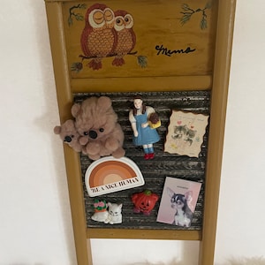 Op de afbeelding: Een houten geframed message board met een zwarte metalen insert. Het bord heeft een geschilderd beeld van uilen en het woord "Mama" bovenaan. De metalen insert heeft een verscheidenheid aan magneten, waaronder een regenboog, een pompoen, een kat, een hond, een teddybeer, een meisje in een blauwe jurk en een kaart met de tekst "Ik hou van je gewoon omdat".