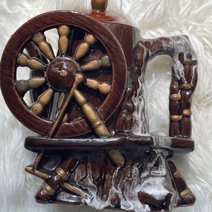 May include: A brown ceramic teapot with a spinning wheel design. The teapot has a handle and a lid. The spinning wheel is made of gold and brown ceramic.