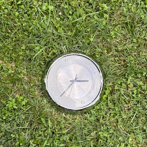 May include: A round, silver-toned clock with a clear face and black hands, resting on a bed of green grass. The clock's face is visible, displaying the time. The clock has a shiny, metallic bezel.