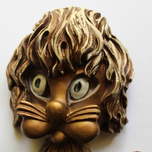 Puede incluir: Una pieza de decoración de pared vintage que presenta una cara caprichosa, de estilo de dibujos animados, con ojos grandes y blancos, una cabeza peluda marrón y blanca y un cuerpo de color dorado.