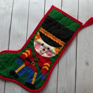 May include: A quilted Christmas stocking featuring a toy soldier design. The stocking has a green background with a diamond pattern, a red border, and a loop for hanging. The toy soldier has a black hat, red jacket, and blue pants.