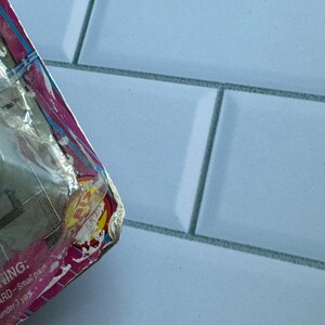 May include: A partially visible, damaged package with pink, yellow, and blue accents. The packaging is wrapped in clear plastic. The background is a light blue tiled surface.