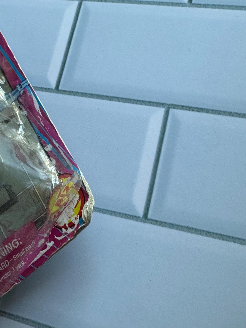 May include: A partially visible, damaged package with pink, yellow, and blue accents. The packaging is wrapped in clear plastic. The background is a light blue tiled surface.