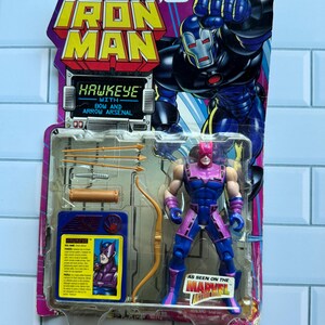 May include: A Hawkeye action figure from the Marvel Iron Man series, packaged on a colorful card. The figure is blue and purple, with a bow and arrow arsenal. The packaging features the Iron Man logo and the text "Hawkeye with Bow and Arrow Arsenal."