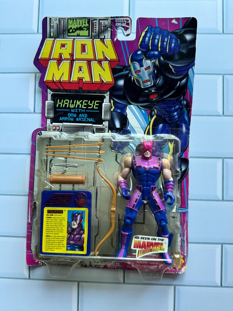 May include: A Hawkeye action figure from the Marvel Iron Man series, packaged on a colorful card. The figure is blue and purple, with a bow and arrow arsenal. The packaging features the Iron Man logo and the text "Hawkeye with Bow and Arrow Arsenal."