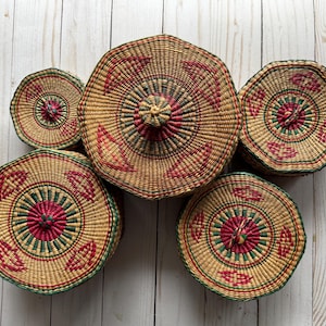 May include: A collection of woven baskets with lids. The baskets are circular, with a natural tan color and accents of red and green. The lids have a central knob and decorative patterns. The baskets are arranged on a light wood surface.