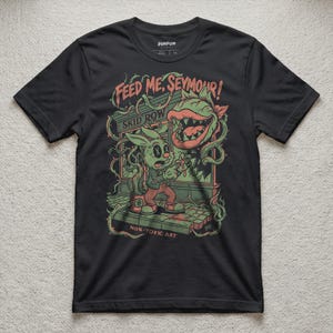 May include: A black short-sleeve t-shirt featuring a graphic illustration of a rabbit and a large carnivorous plant. The text reads "FEED ME, SEYMOUR!", "SKID ROW", and "NON-TOXIC ART". The design uses green, red, and white colors on a dark background.