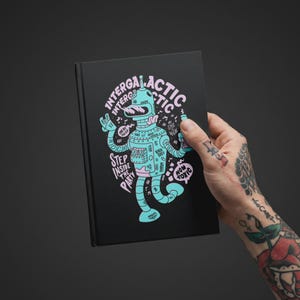 May include: A black hardcover notebook featuring a light blue and pink graphic illustration of a robot. The text reads INTERGALACTIC, STEP INSIDE THE PARTY, MY STYLE IS WILD, and NON TOXIC, surrounded by small musical notes and various decorative symbols.