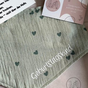 May include: A light green bandana with dark green heart patterns and the word "Geburtstagskind" in white script. The bandana is accompanied by two cards, one with care instructions and the other with a logo and text.