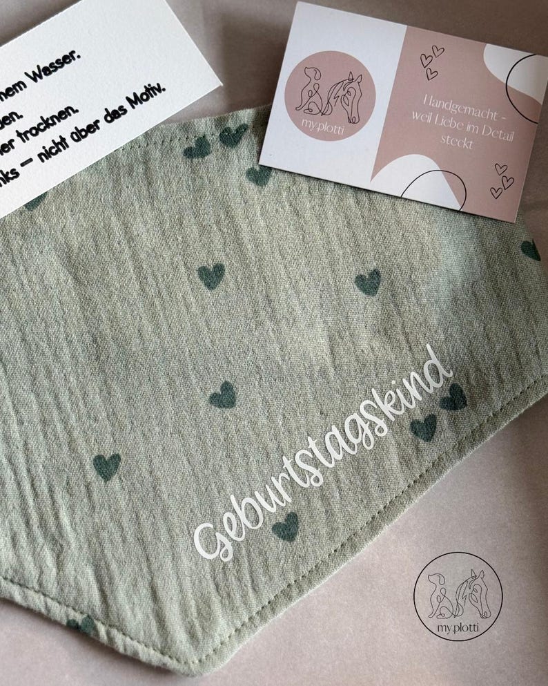 May include: A light green bandana with dark green heart patterns and the word "Geburtstagskind" in white script. The bandana is accompanied by two cards, one with care instructions and the other with a logo and text.