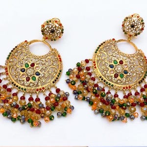 May include: A pair of gold-toned chandelier earrings with intricate filigree detailing. The earrings feature a crescent-shaped design adorned with colorful gemstones and dangling beaded strands in shades of red, green, orange, and yellow.