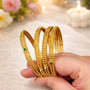 May include: A set of gold bangles with intricate floral and dot patterns. Each bangle features small green and white gemstones. The bangles are held in a hand, showcasing their design and craftsmanship. The background is softly lit with flowers and a candle.