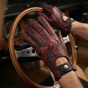 Può includere: Un paio di guanti da guida con un motivo a pelle di serpente testurizzato rosso e nero. I guanti presentano fori circolari sulle nocche, un'apertura sul polso e un cinturino in pelle nera fissato con un bottone a pressione in metallo argentato.
