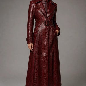 May include: A long, deep red trench coat featuring a textured snakeskin pattern, a wide collar, and a matching belt with a buckle at the waist. The coat has long sleeves with buckled cuffs, side pockets, and a dark inner layer with a front zipper.