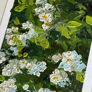 May include: Close-up of an oil painting featuring white flowers with yellow centers and light blue accents, set against a backdrop of lush green foliage. The textured brushstrokes create a three-dimensional effect, capturing the essence of a floral garden.