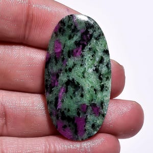 Puede incluir: Gema ovalada con una mezcla de colores verde, negro y magenta. La piedra pulida presenta un aspecto natural y texturado. La gema se sostiene en una mano.