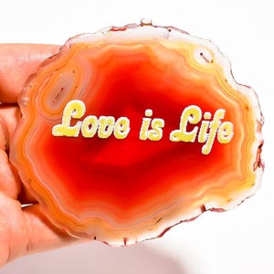 Può includere: Una fetta di agata lucida e traslucida con bande concentriche di vivaci colori arancione, rosso e bianco. Il centro della pietra mostra il testo "Love is Life" in un carattere giallo, grassetto e senza grazie, che contrasta con lo sfondo.