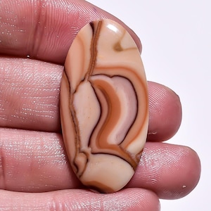 May include: An oval gemstone with a polished surface. The stone displays a swirling pattern of tan, brown, and cream colours. The gemstone is held between fingers, showing its size and shape.