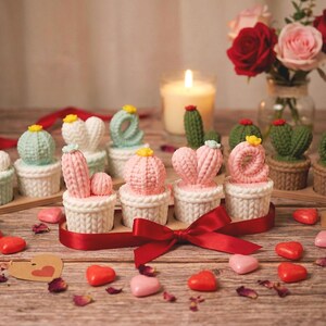 Puede incluir: Una colección de velas con forma de cactus en varios colores, incluyendo rosa, verde y blanco, dispuestas sobre una superficie de madera. Las velas están en pequeñas macetas de estilo tejido. Caramelos rojos en forma de corazón y una cinta roja completan la decoración.