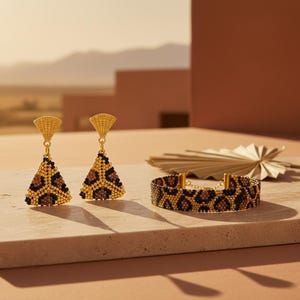 Könnte beinhalten: Goldfarbenes Schmuckset mit dreieckigen Ohrringen und einem Armband, beide mit Leopardenmuster. Die Ohrringe haben einen fächerförmigen Aufsatz. Das Armband hat einen goldenen Kettenverschluss. Präsentiert auf einer neutralen Oberfläche, mit Wüstenhintergrund.