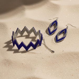 Può includere: Un set di gioielli di perline con un braccialetto e orecchini. Il braccialetto e gli orecchini hanno un motivo a zig-zag con perline blu e argento. Gli orecchini sono a forma di goccia. I gioielli sono esposti su una superficie sabbiosa.