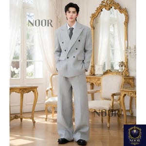 May include: A light gray double-breasted suit with wide-leg pants. The suit includes a matching tie and a white dress shirt. The word "NOOR" is printed on the top left of the image. The suit is worn with black dress shoes.