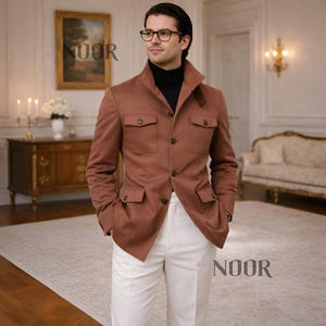 Puede incluir: Una chaqueta de color óxido con cuatro bolsillos delanteros y cierre de botones. La chaqueta se combina con un cuello alto negro y pantalones blancos. La imagen también muestra a un hombre con gafas.