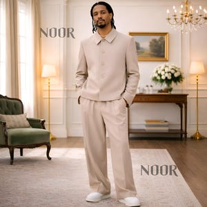 May include: A beige suit with a button-up jacket and matching pants. The suit is worn with white sneakers. The word "NOOR" is printed in the upper left and lower right corners of the image. The suit is a neutral color.