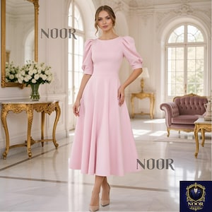Puede incluir: Un vestido midi rosa claro con mangas cortas abullonadas y un corpiño ajustado. El vestido tiene una falda acampanada y se usa con tacones nude. La palabra "NOOR" se muestra en la imagen.
