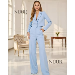 Puede incluir: Un traje de dos piezas azul claro con una chaqueta corta y pantalones de pernera ancha. La chaqueta tiene un cierre de un solo botón y solapas con muescas. Los pantalones tienen cintura alta y bolsillos. La palabra "NOOR" está impresa en las esquinas superior izquierda e inferior derecha.