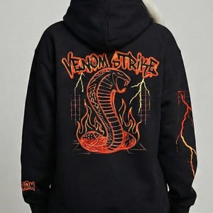 May include: Black hoodie featuring a cobra design with the text "Venom Strike" in orange and yellow. The cobra is surrounded by flames and lightning bolts. The hoodie has a hood and long sleeves.