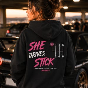 Op de afbeelding: Zwarte hoodie met de tekst "SHE DRIVES STICK" in roze en wit, en een versnellingsbakdiagram. De zin "THREE PEDALS. TOTAL CONTROL." staat eronder. De hoodie wordt door een persoon gedragen.