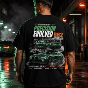 Op de afbeelding: Een zwart t-shirt met een grafische print van een groene sportwagen. De tekst luidt PRECISION EVOLVED G82, COMPETITION xDRIVE, GERMAN PRECISION, ALL-WHEEL POWER, TWIN-TURBO, 503 HP en xDRIVE. Het ontwerp toont de auto vanuit voor- en achteraanzicht.